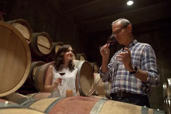 Dues persones tastant vi negre entre bótes de roure al celler